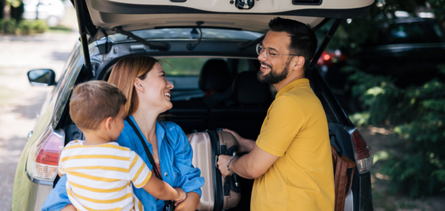 A happy family of three is seen smiling and chatting while packing their car trunk for a family road trip.