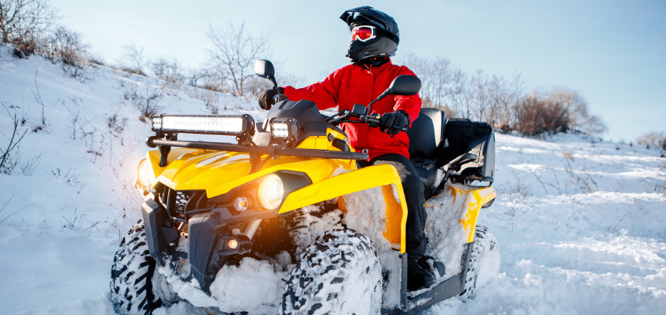 A person in a red jacket and black helmet rides a bright yellow ATV through deep snow on a sunny winter day with ATV insurance.