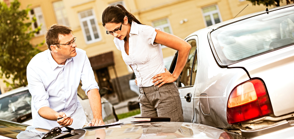 Two drivers looking frustrated while filling out an accident report after a staged collision in a parking lot.