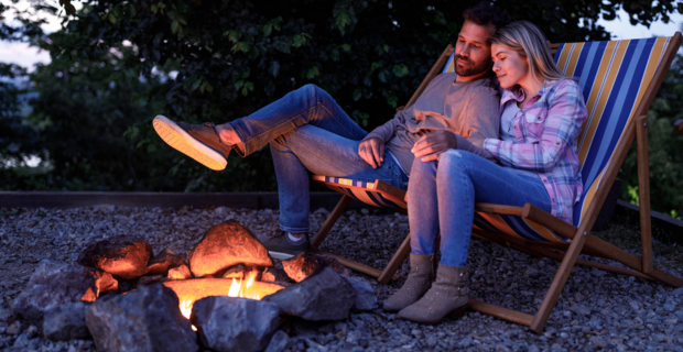 A man and a woman hold hands as they sit by an outdoor fire on a quiet evening.