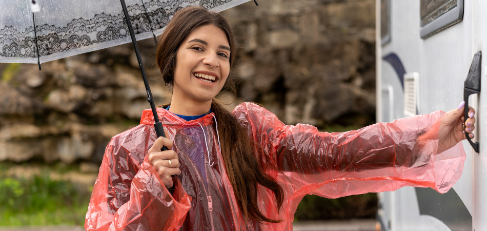 A woman seen smiling with an umbrella and rain poncho about to open the door to her motorhome.