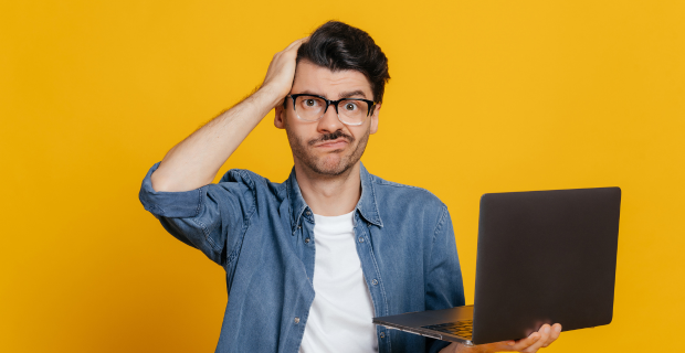 Young adult holding his laptop in one hand and his head in the other standing upright with a confused look on his face in front of an orange background.