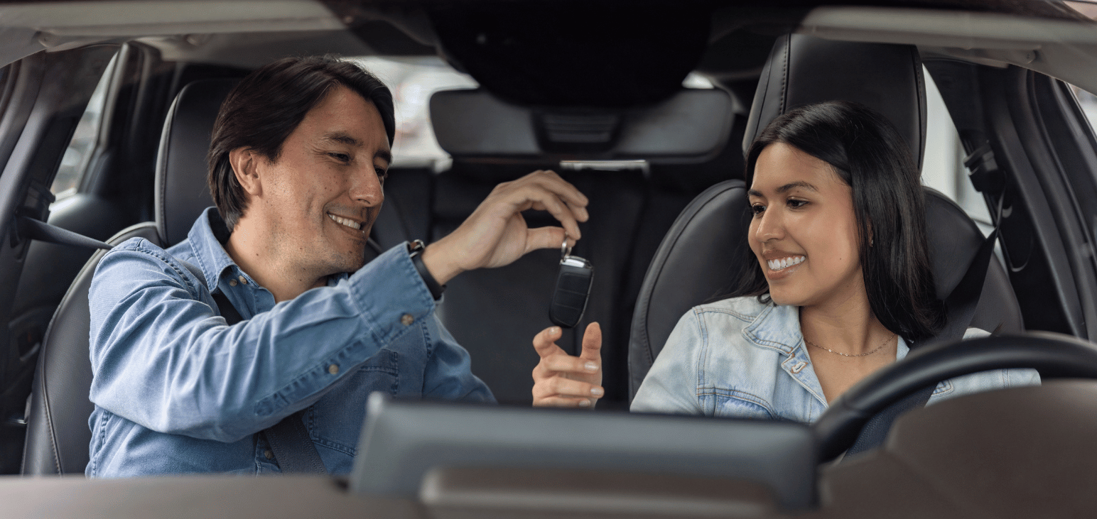 A father hands over the keys to his daughter in a vehicle. 