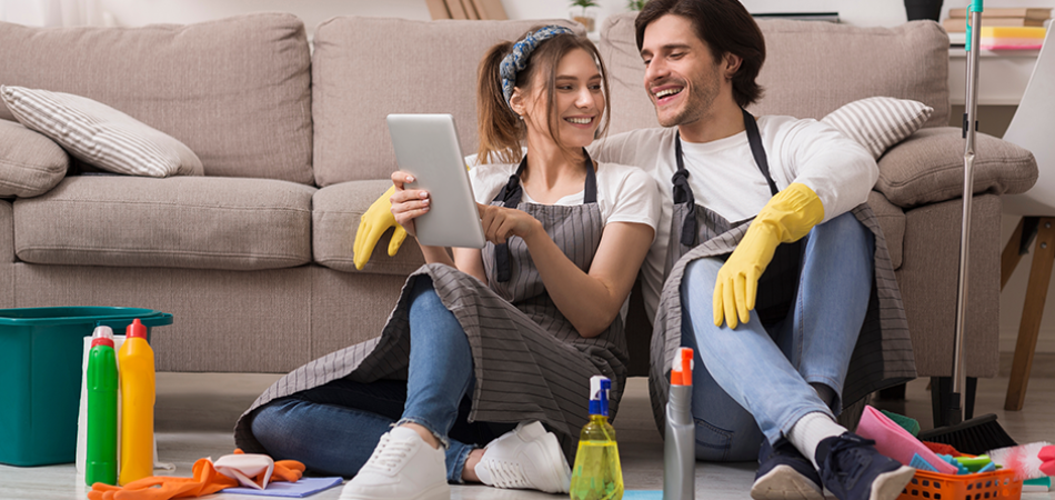 Two people in aprons sit on the floor surrounded by cleaning supplies while looking at a tablet.