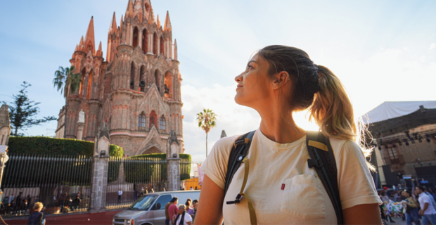 A women standing near a busy street in Mexico on a sunny day. There is a tall building in the background.