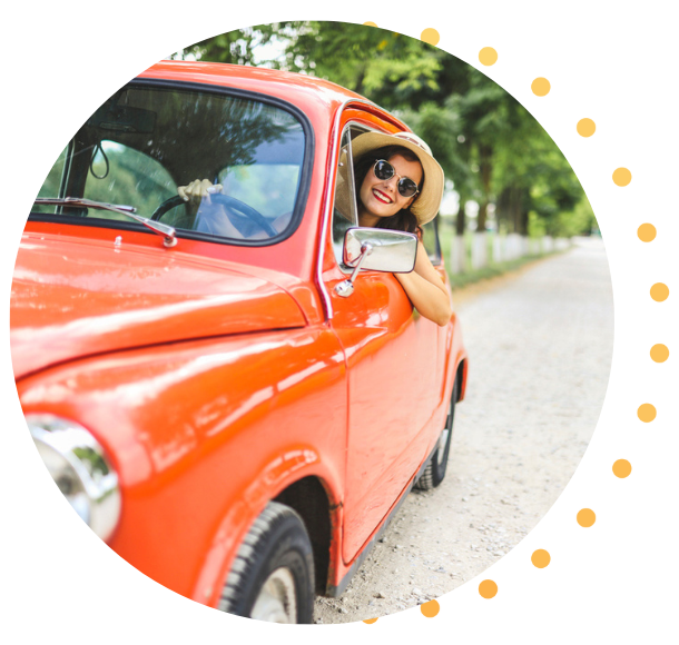 A woman in a sunhat leans out of the driver's side window of her bright orange classic car.