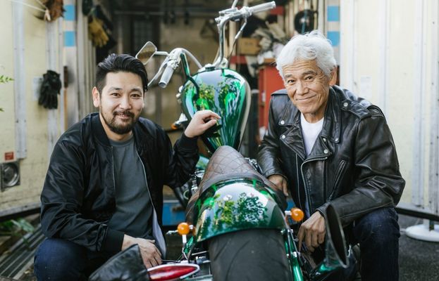 An Asian man in a bomber jacket and his grandfather smile, posing on either side of a cruiser. 