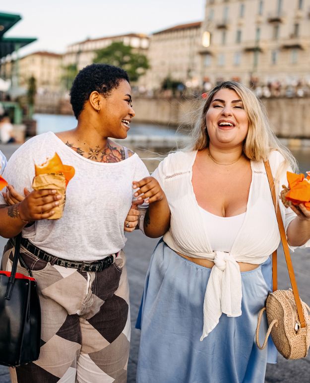 Two friends walk along the canal, laughing and enjoying local delicacies while on vacation. 