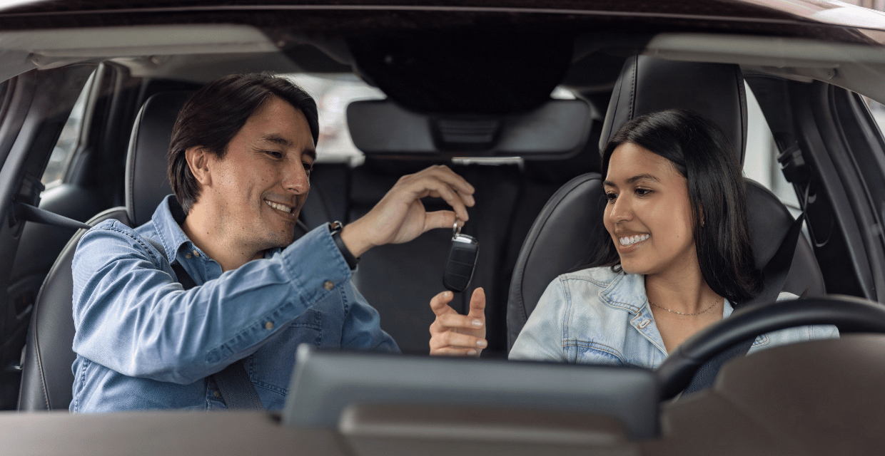 A father hands over the keys to his daughter in a vehicle. 