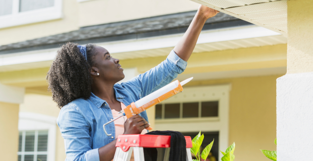 A woman is seen on a ladder next to her house on a sunny day with a caulking gun sealing here roof to protect against flooding.