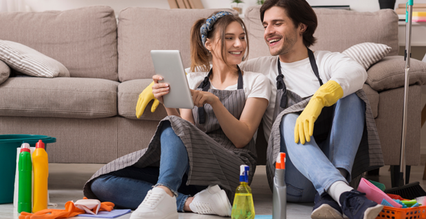 Two people in aprons sit on the floor surrounded by cleaning supplies while looking at a tablet.